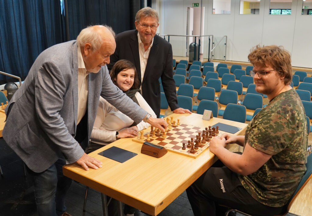 Rüdiger Meier, der Bürgermeister der Gemeinde Kirchlengern, führte den ersten Zug am Brett der Titelverteidiger*innen Marla Windmüller von der SG Hücker-Aschen und Matts Struckmeier von der SG Bünde aus. Hinter Windmüller der Bezirksvorsitzende Hermann Dieckmann.
